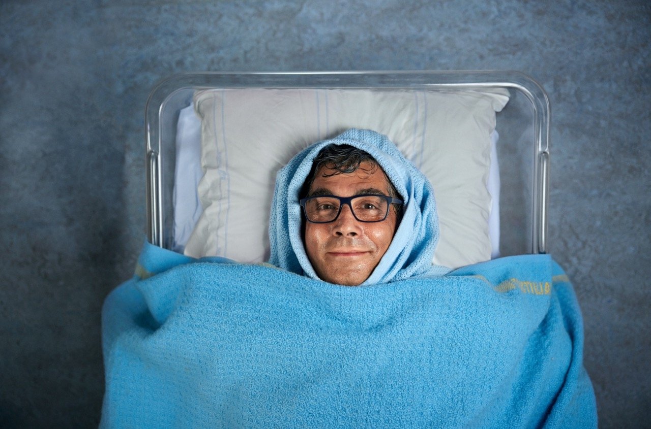 En man i 60-årsåldern med glasögon som ligger insvept i en blå filt i en plastbalja, det ser ut som att han är nyfödd.